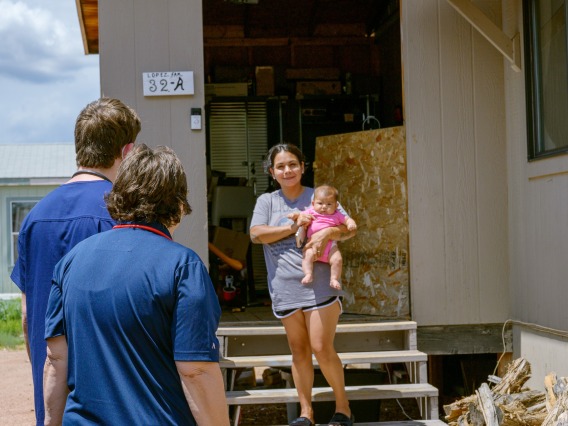A photograph of a mom walking out of her house holding a baby. She is being met by two health professionals wearing stethoscopes and blue scrubs.